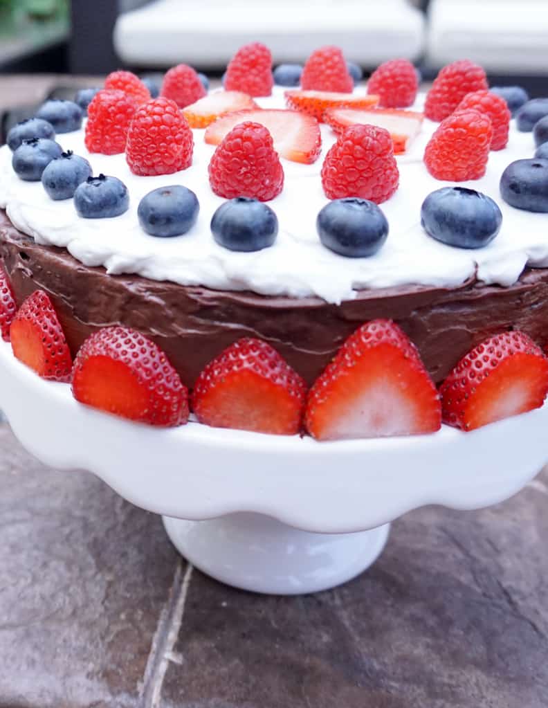 Vegan chocolate sweet ptoato cake on white cake stand with strawberry border and mixed berries on top for a red white and blue celebration.