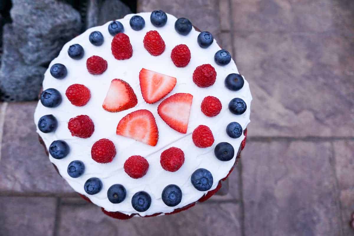strawberries, raspberries and blue berries aranged in concentric circles on coconut whip cream on a chocolate cake near a fire pit.
