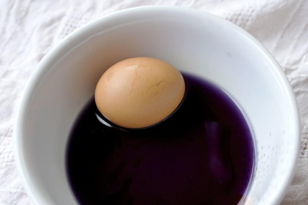 A brown egg in purple dye in a white bowl