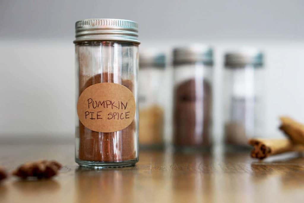 Glass jar of pumpkin pie spice mix on wooden table with other spices around