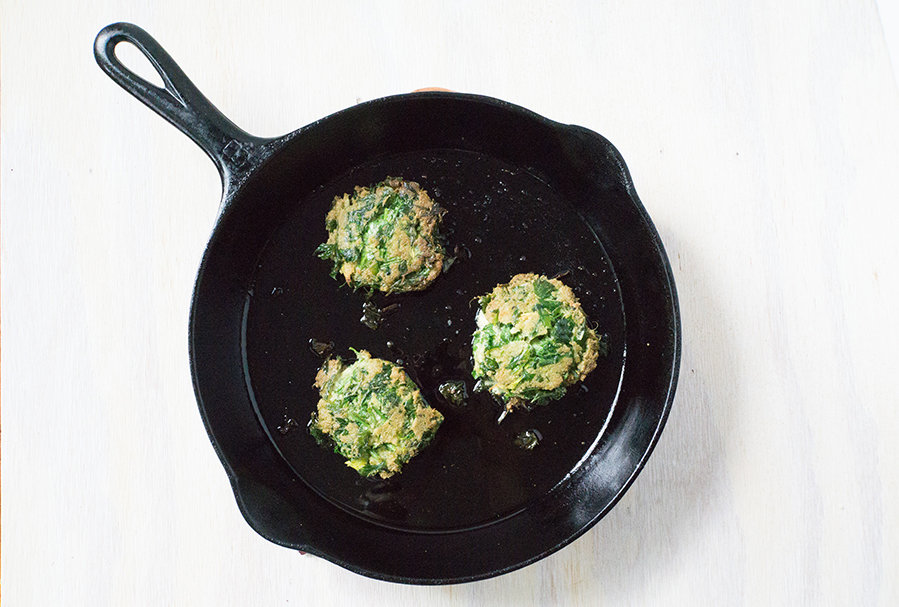 3 celery fritters frying in a cast iron skillet on white wood background