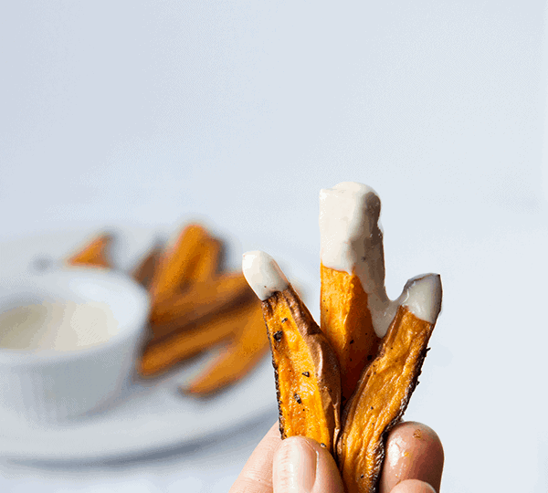 hand holding 3 sweet potato fries dipped in whipped tahini sauce with sauce and fries in the background.