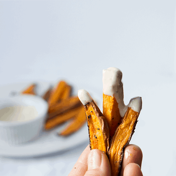 hand holding 3 sweet potato fries dipped in whipped tahini sauce with sauce and fries in the background.