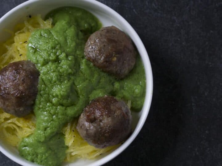 Green sauce smothered meatballs on spaghetti squash in white bowl on dark marble and poured concrete surface
