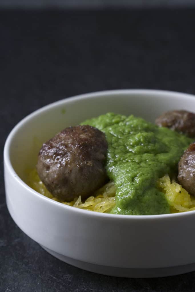 Green sauce smothered meatballs on spaghetti squash in white bowl on dark marble and poured concrete surface