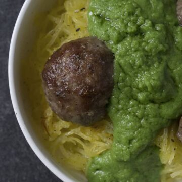 Green sauce smothered meatballs on spaghetti squash in white bowl on dark marble and poured concrete surface