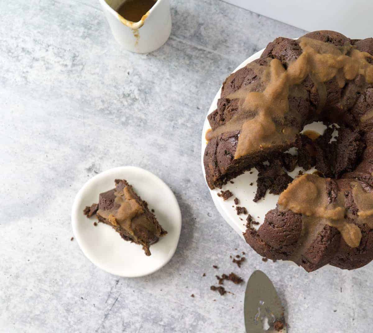 Coconut cardamom glazed chocolate chai bundt cake on white cake stand on cement table with white backdrop with slice on plate, server, and jar of glaze on table.
