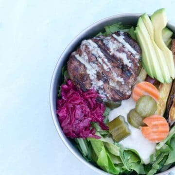Paleo Elk Burger Bowl in Tin bowl on cement background: mixed greens topped with cashew cream, pickled cucumbers and carrots, roasted sweet potato fries, red sauerkraut, elk burger and tahini