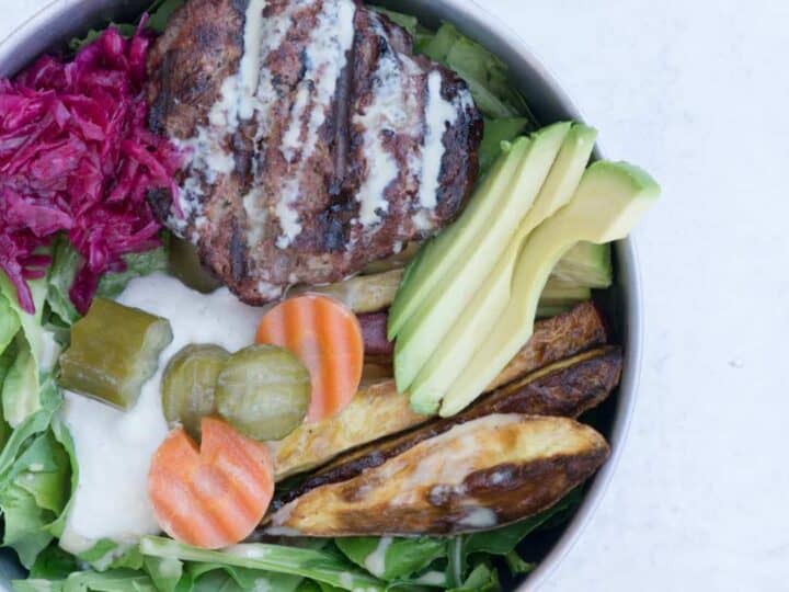 Paleo Elk Burger Bowl in Tin bowl on cement background: mixed greens topped with cashew cream, pickled cucumbers and carrots, roasted sweet potato fries, red sauerkraut, elk burger and tahini