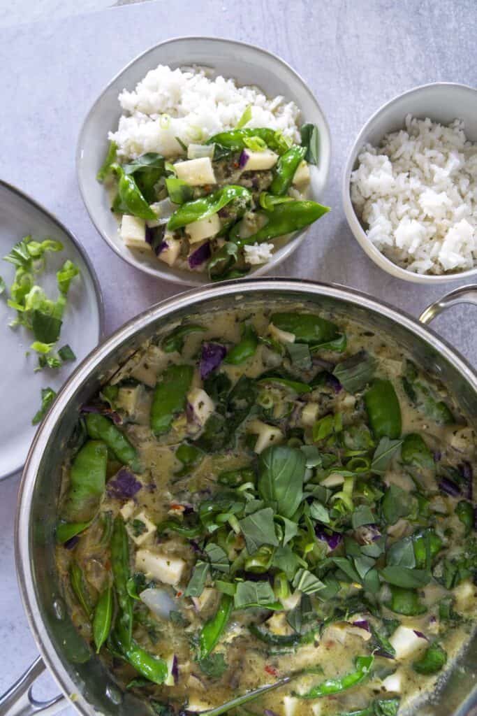 Snap pea and johlrabi curry in a pot with bowl of rice and green onions for garnish